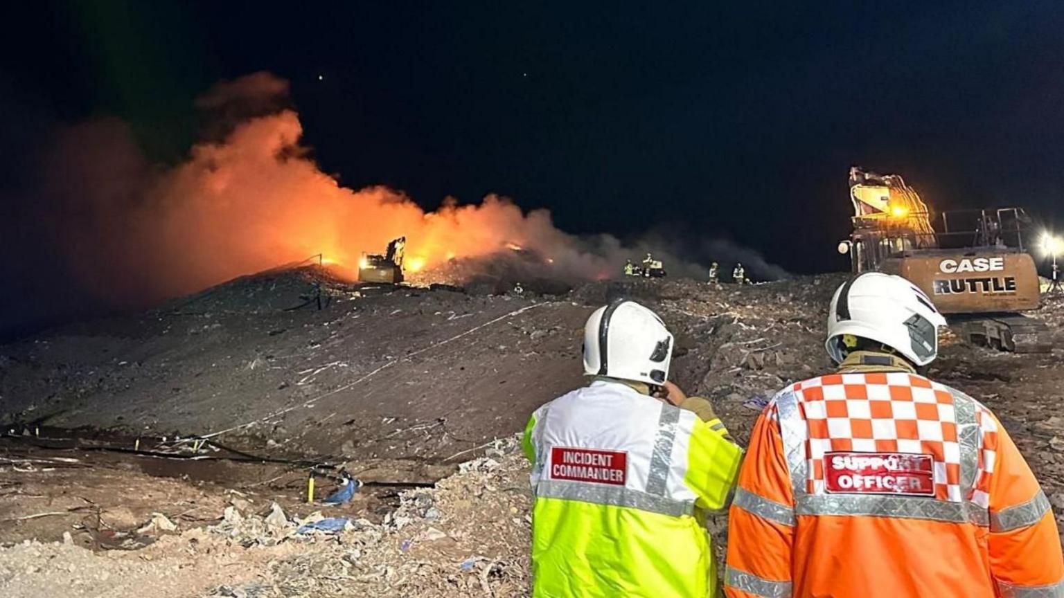 Two men in white helmets and hi-vis jackets in the foreground watch a fire at a landfill