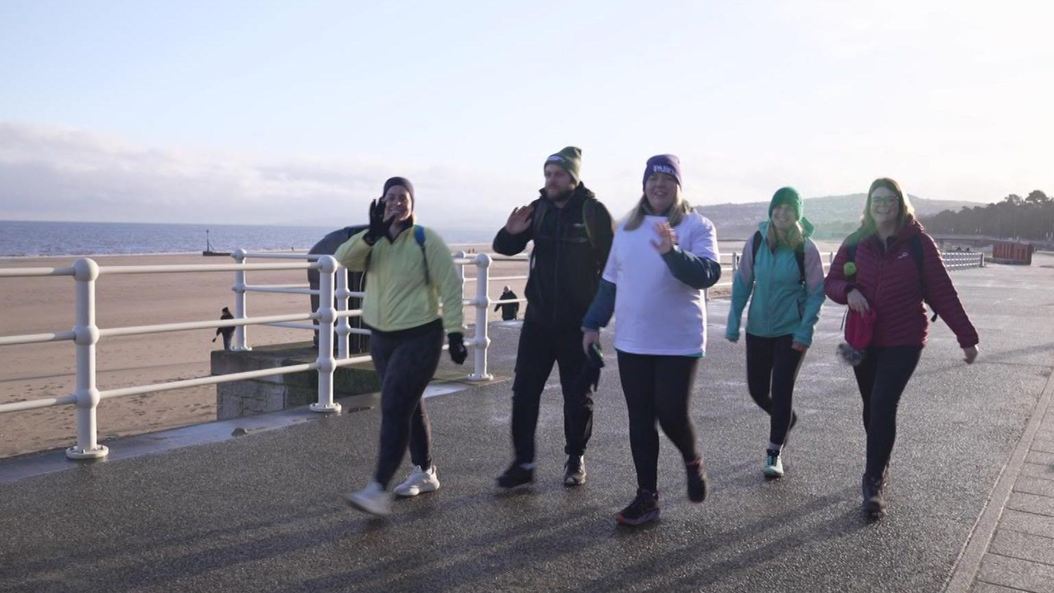 Pedair menyw ac un dyn yn cerdded ar hyd prom rhwng Bae Colwyn a Chonwy gan godi llaw at y camera