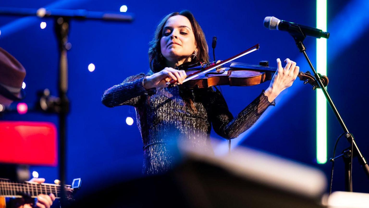 A fiddler plays her instrument as she stands next to a microphone and against a blue backdrop.