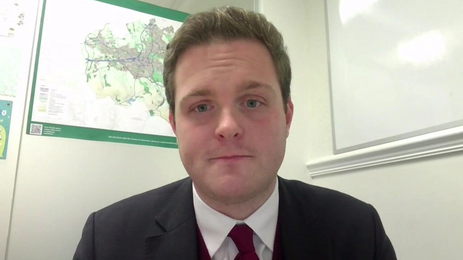 Oliver Ryan on a Teams call in his office. He is wearing a dark suit, white shirt and dark red tie. He has a clean shaven face and light brown hair.