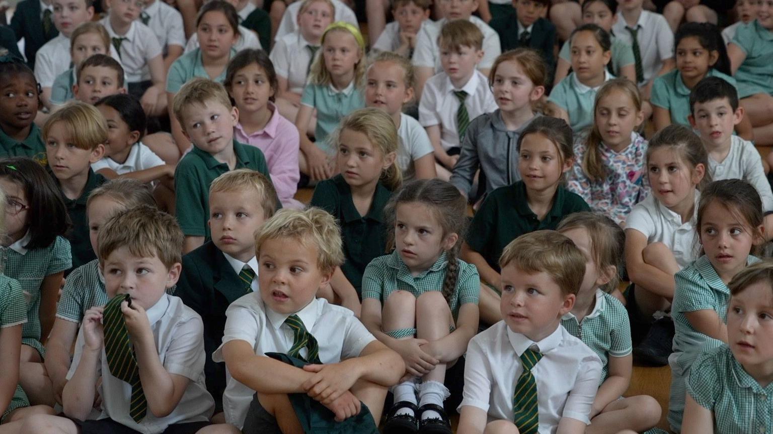 Primary school pupils sit and listen, some with quizzical expressions.