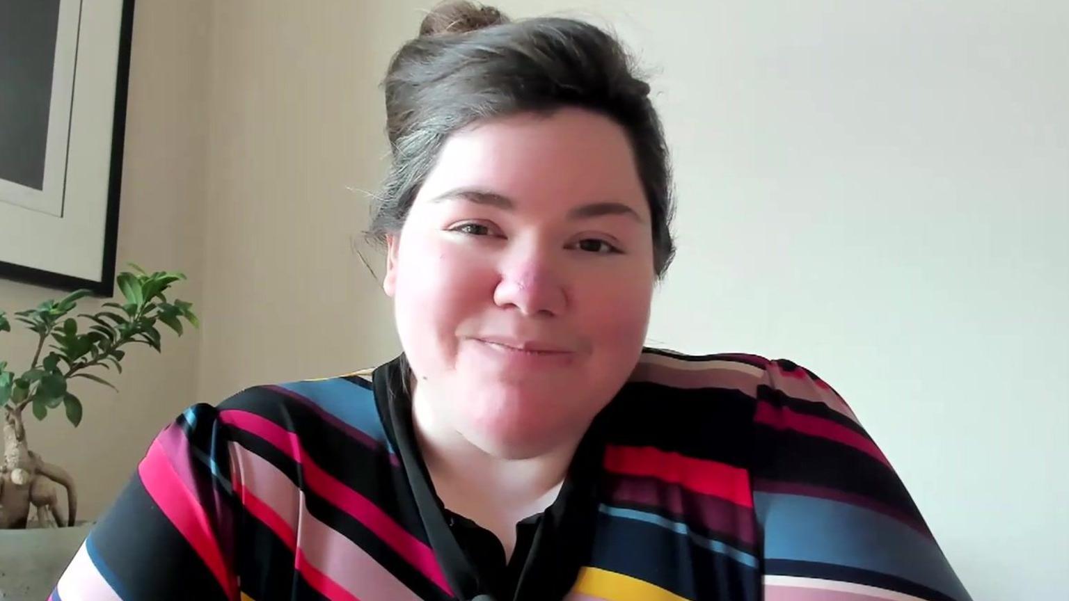 Rachel Clarke with her brown hair tied up in a bun. She is wearing a black blouse with multi-coloured, diagonal stripes on it