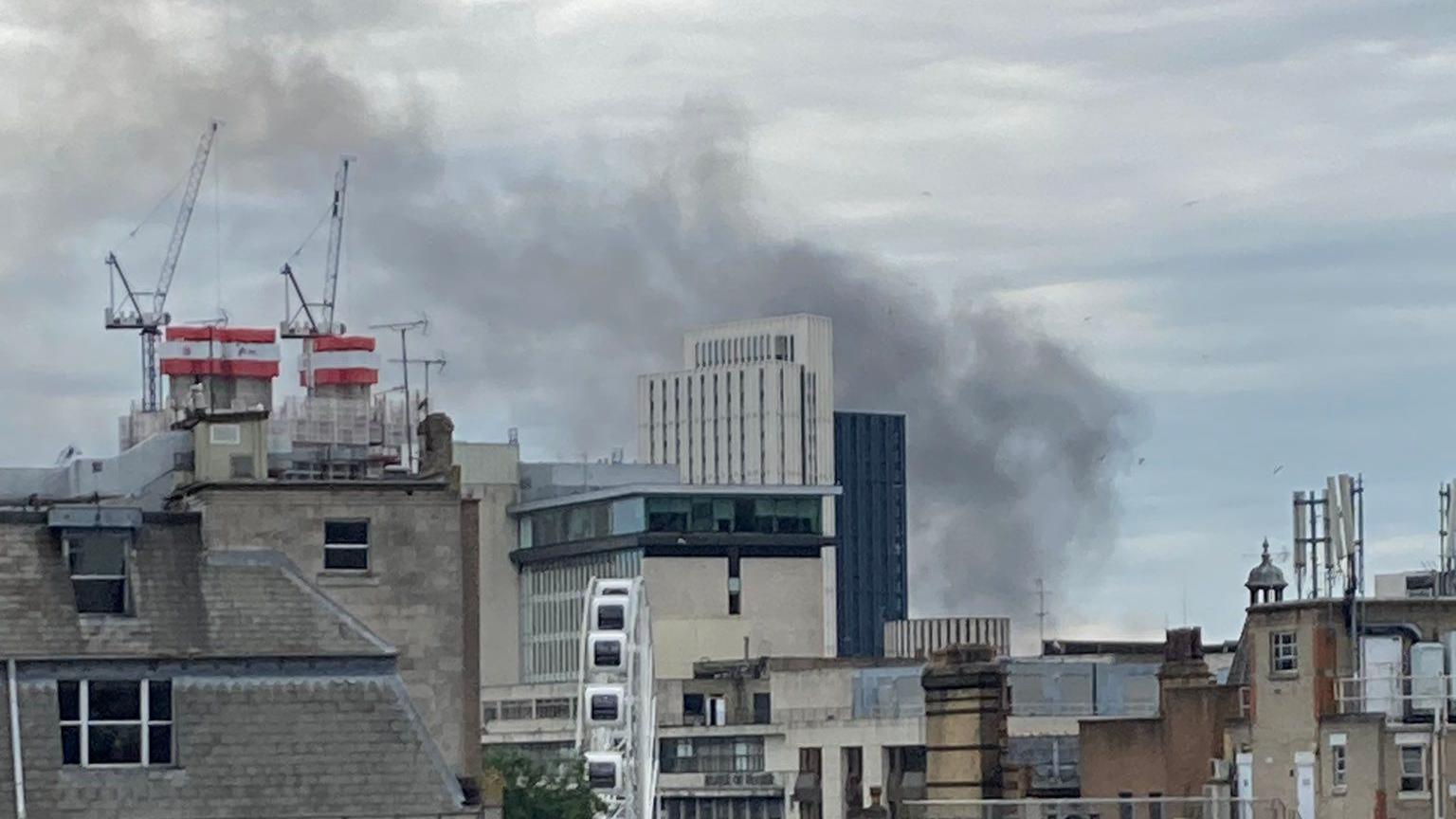 Smoke billowing into the air from a building, various other buildings are visible along the skyline