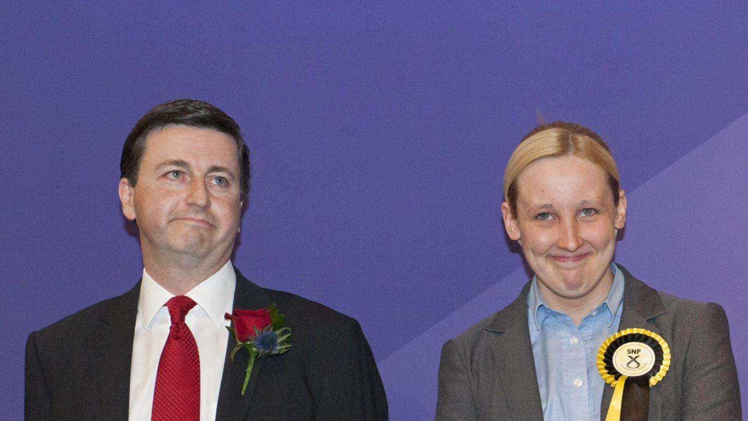 The election count in 2015 - Douglas Alexander -  a dark haired man with a suit, red tie and red rosette, stands next to Mhairi Black, who has  blonde hair and is wearing a grey suit and an SNP rosette