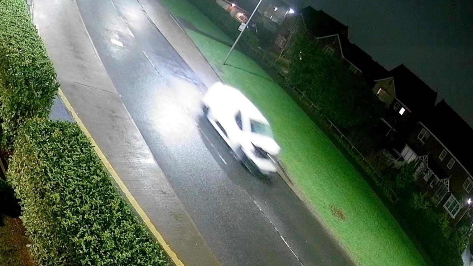 Still from a CCTV footage of a white van driving on a road past houses, its windscreen is smashed and bonnet is crumpled upwards.