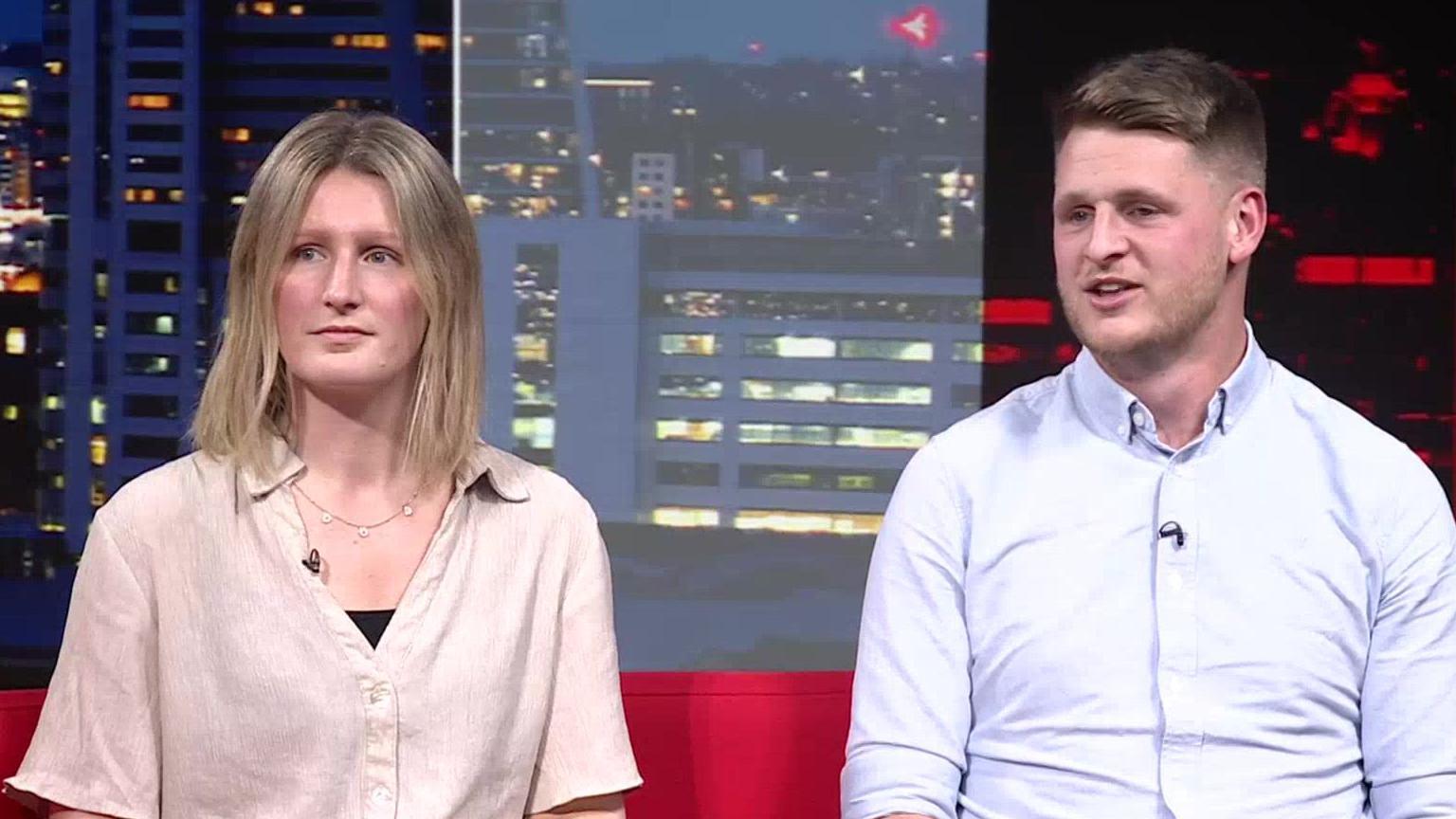 Laura and Ben are seated on a bright red couch in a studio setting. Laura is wearing a light beige short-sleeved shirt over a black inner layer and has a necklace. Ben is wearing a light blue button-up shirt with rolled-up sleeves. Behind them is a large backdrop showing a cityscape at night with illuminated buildings.