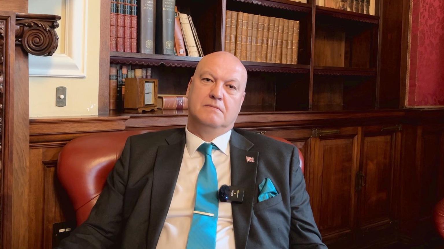 Ian Cooper wears a turquoise blue tie, a suit jacket, and is sat in an armchair frowning.