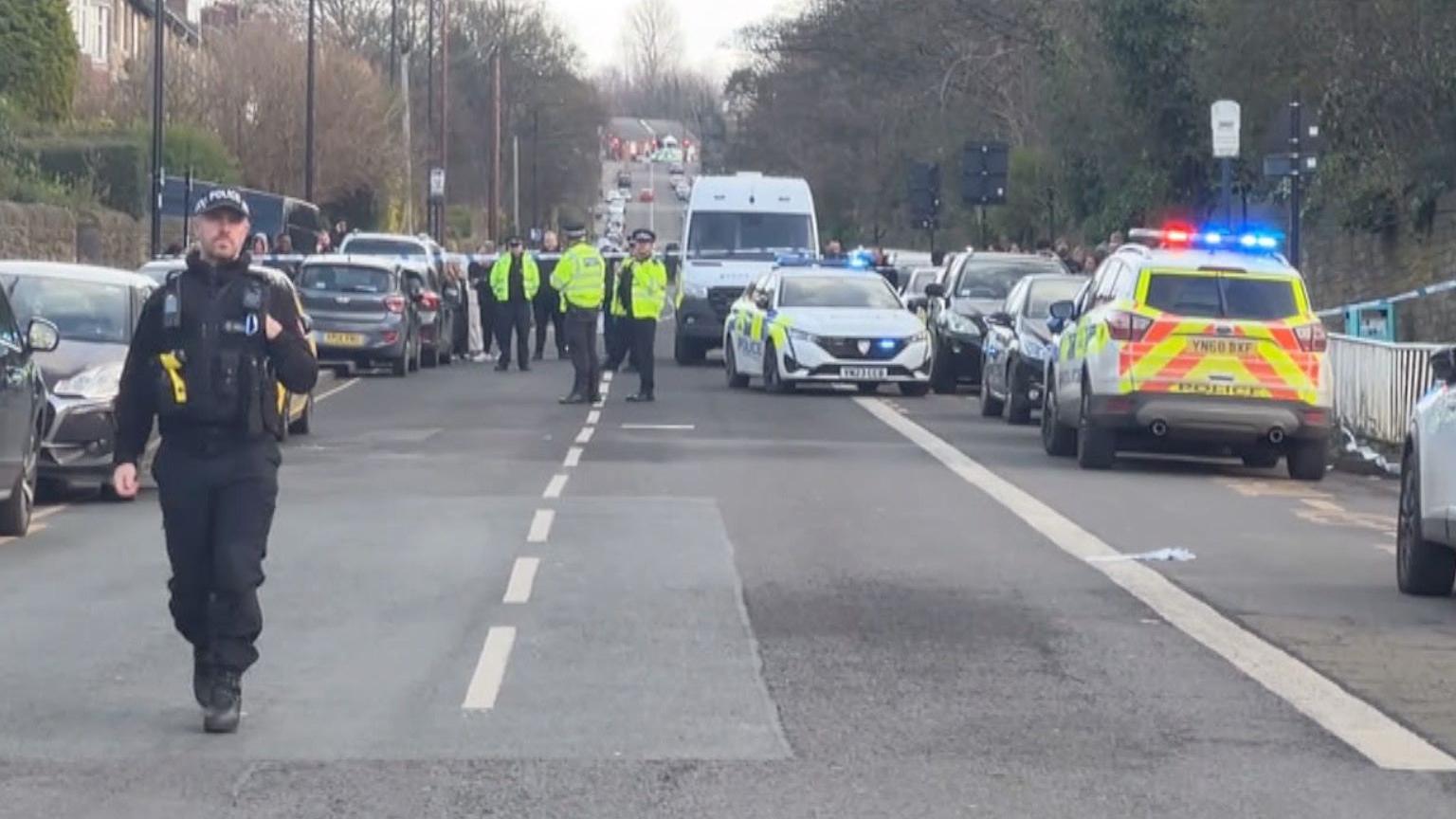 Police officer approaches camera as numerous emergency vehicles line eaither side of street strung with crime scene tape