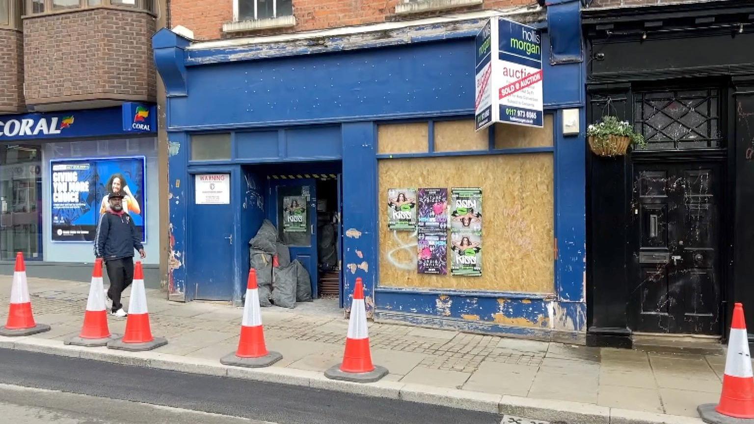 An empty shop is the primary image of this photo. The shop has been boarded up with posters on the boards. The door is open and there's a number of bin sacks in the porch. There's a person walking past the shop looking at the camera. Red and white cones are on the pavement in front of the shop and there's a coral bet shop on the left hand side of the image. At the front of the shop there's also a sign, saying 'sold at auction' from an estate agent.