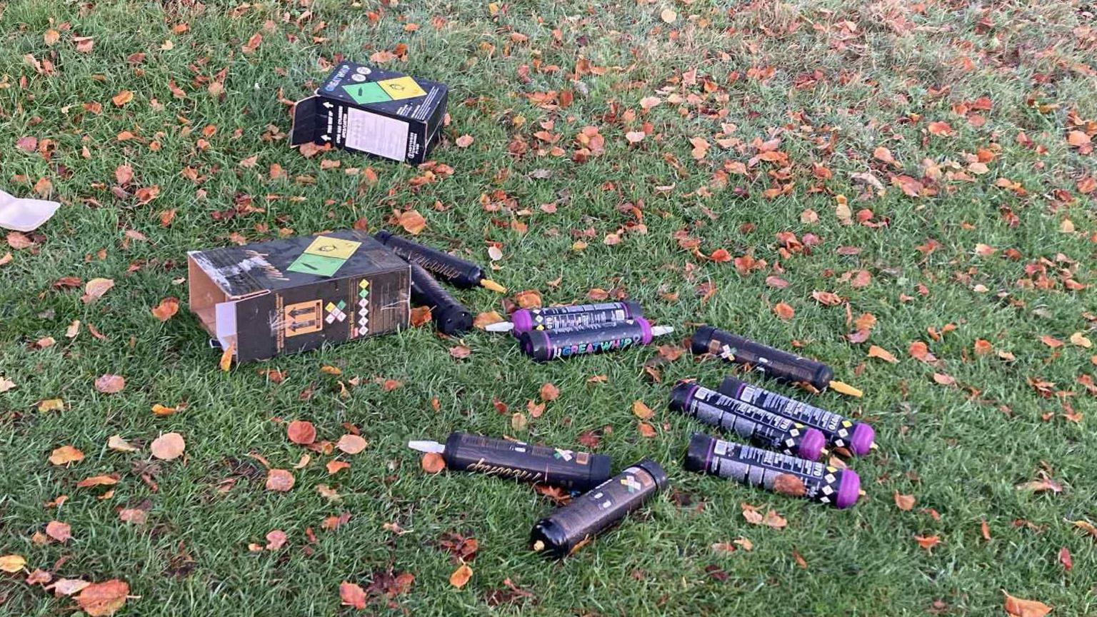 A patch of autumnal grass with scattered brown leaves resting on it. There are also about a dozen large canisters with danger warnings printed all over the packaging. Two boxes are also littered with small balloons weaved into the grass. 