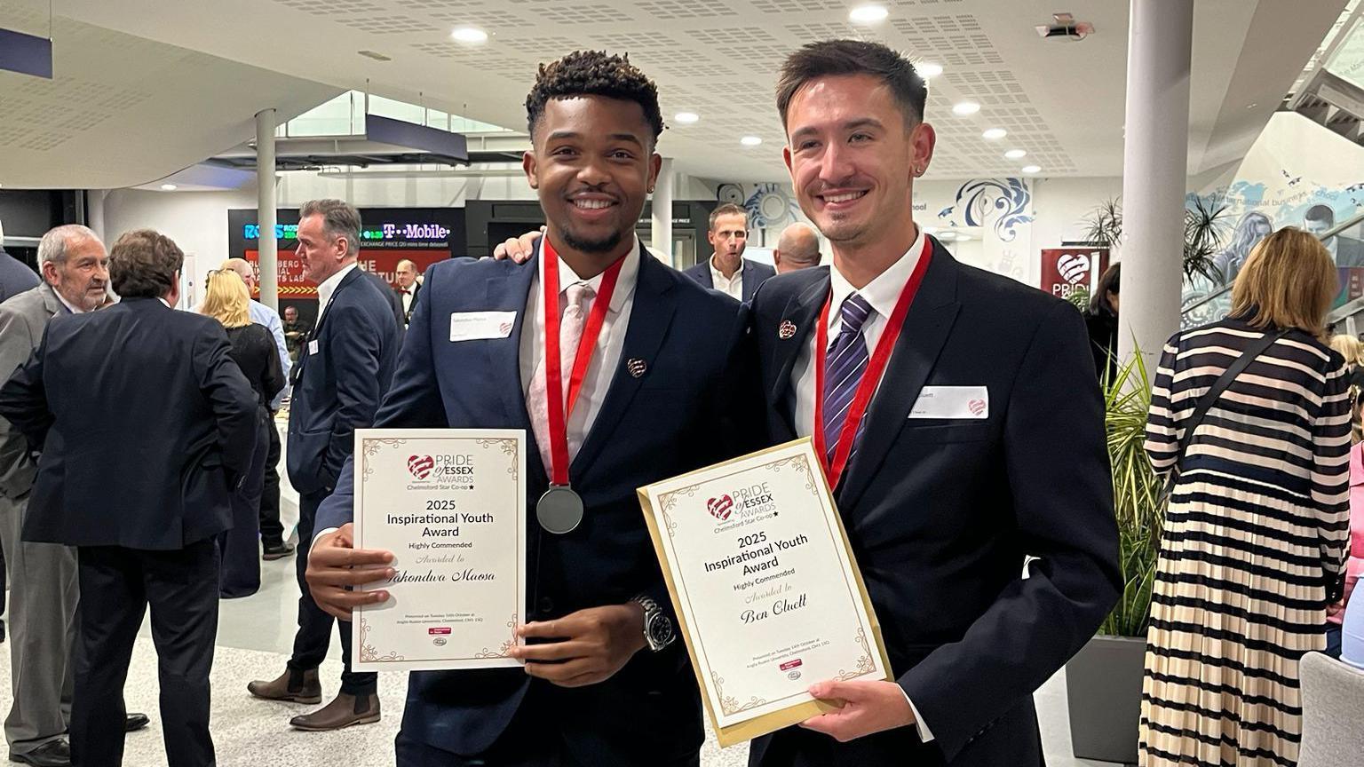 Takondwa Maosa and Ben Cluett in navy blue suits, white shirts and ties standing in a foyer area, both holding certificates, and with red-ribbonned medals around their necks. They are smiling and Ben has his arm around Takondwa.