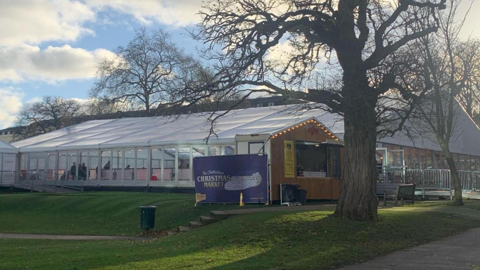 The ice rink in Imperial Gardens, Cheltenham. It is a large white marquee on a grassy field with festoon lighting and an advertisement sign for the Christmas Market.