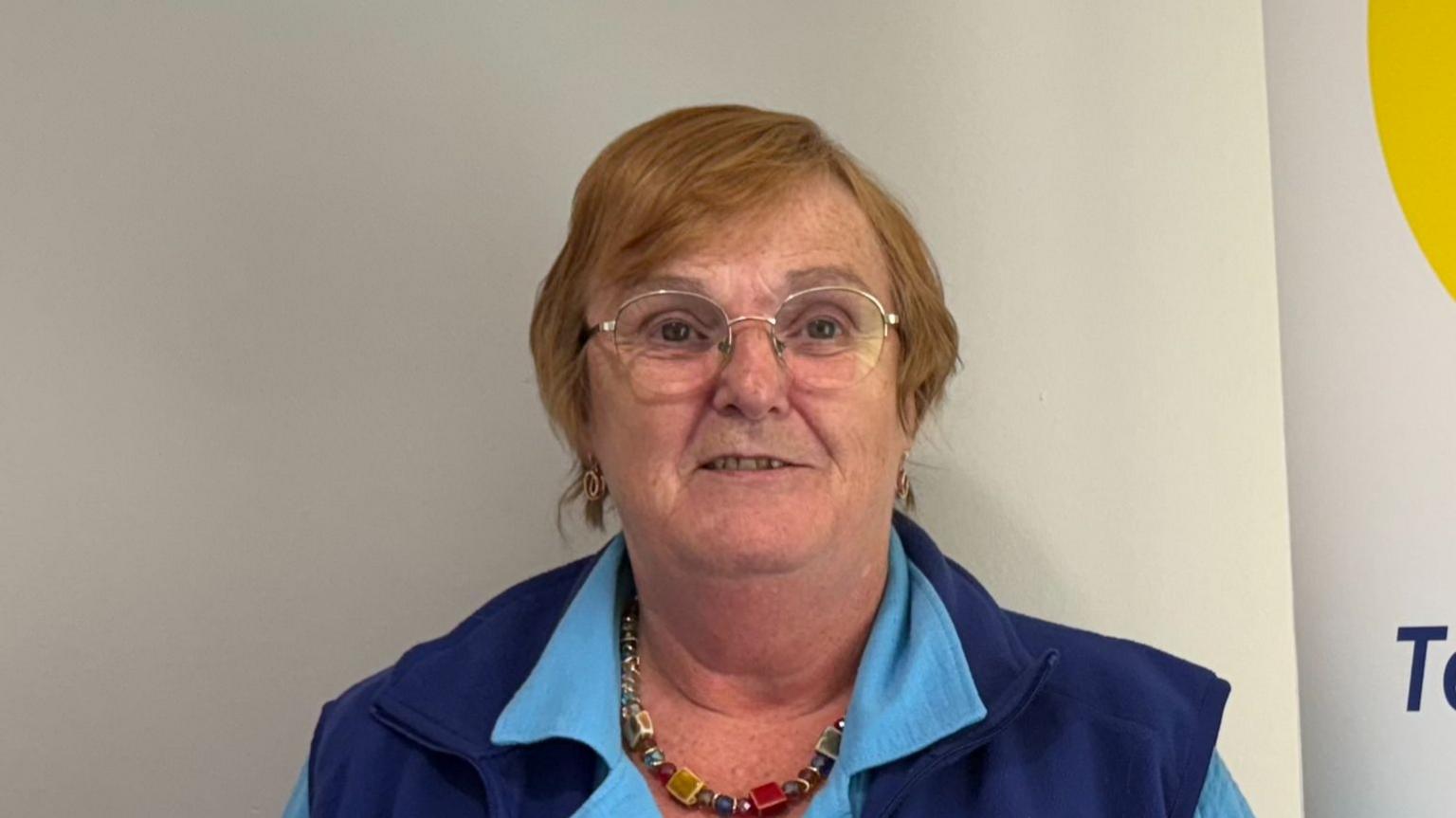 A woman with short light coloured hair is looking into the camera. She is metal rimmed glasses, a light blue polo neck top and a dark blue sleeveless gilet.