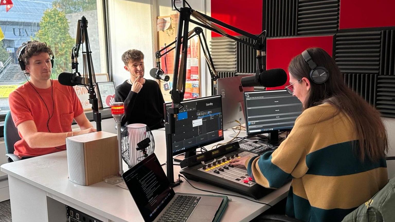 Greg James is sitting behind a radio desk next to Nathan Winterford. On the other side of the desk, and whose back is towards the camera, is Max Gowers.
