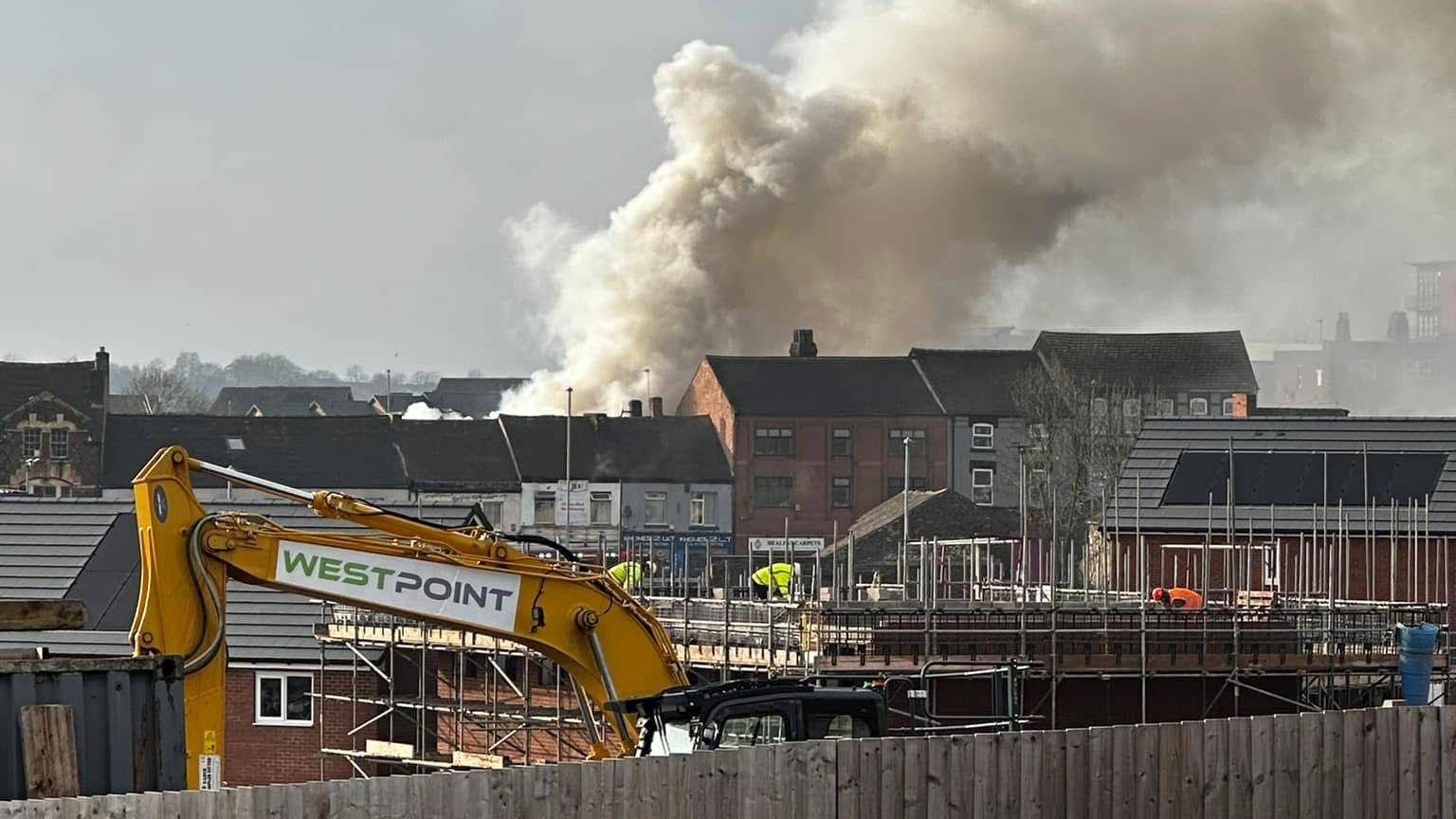 A large plume of smoke above a row of houses. There is a digger in the foreground and construction workers on a building site.