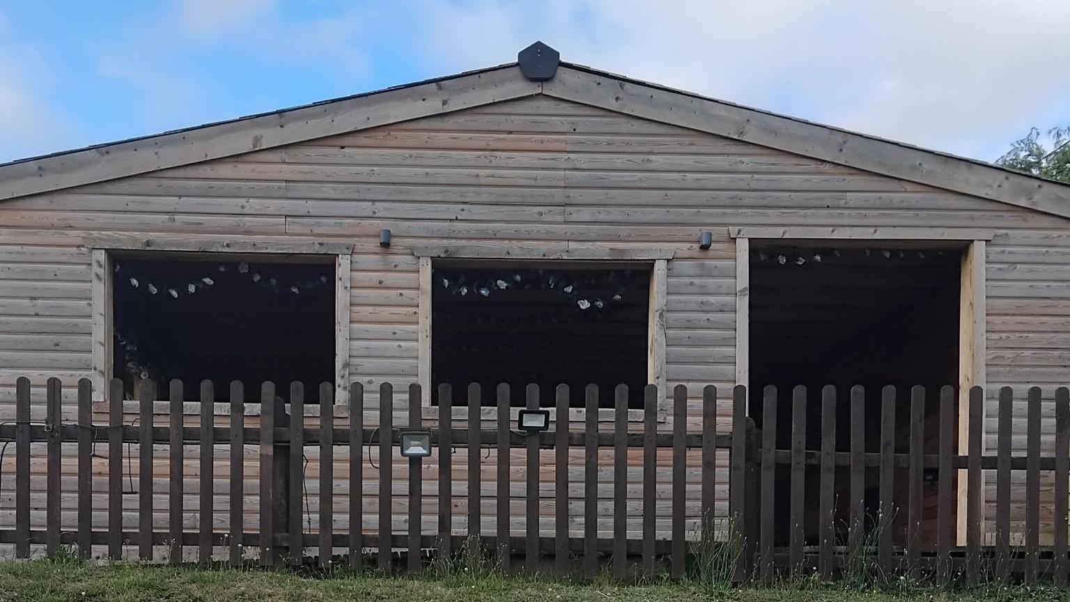 Outside of large wooden outdoor seating shed behind a brown wooden fence. The shed has two large open windows and a open doorway. There are two flood lights attached to the fence.