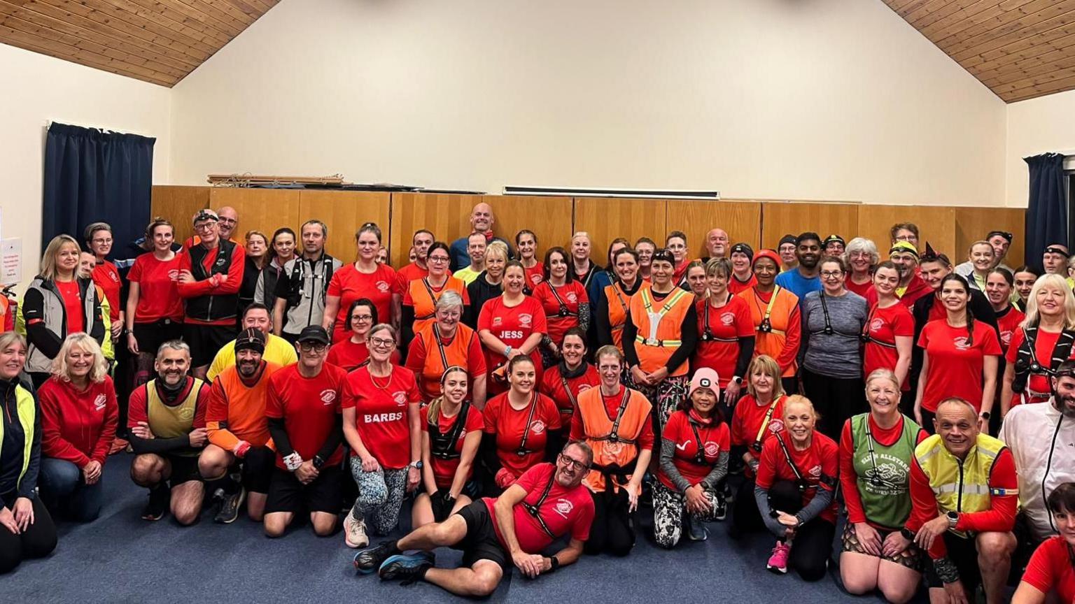 A large group of about 50 people gathered inside a hall with a high, wooden, vaulted ceiling and fluorescent strip lighting. Most are wearing bright running attire in shades of red, orange, and yellow, with reflective straps and head torches. The group is arranged in rows, some kneeling at the front and others standing behind, smiling towards the camera. The hall has cream walls and dark blue curtains on the windows.