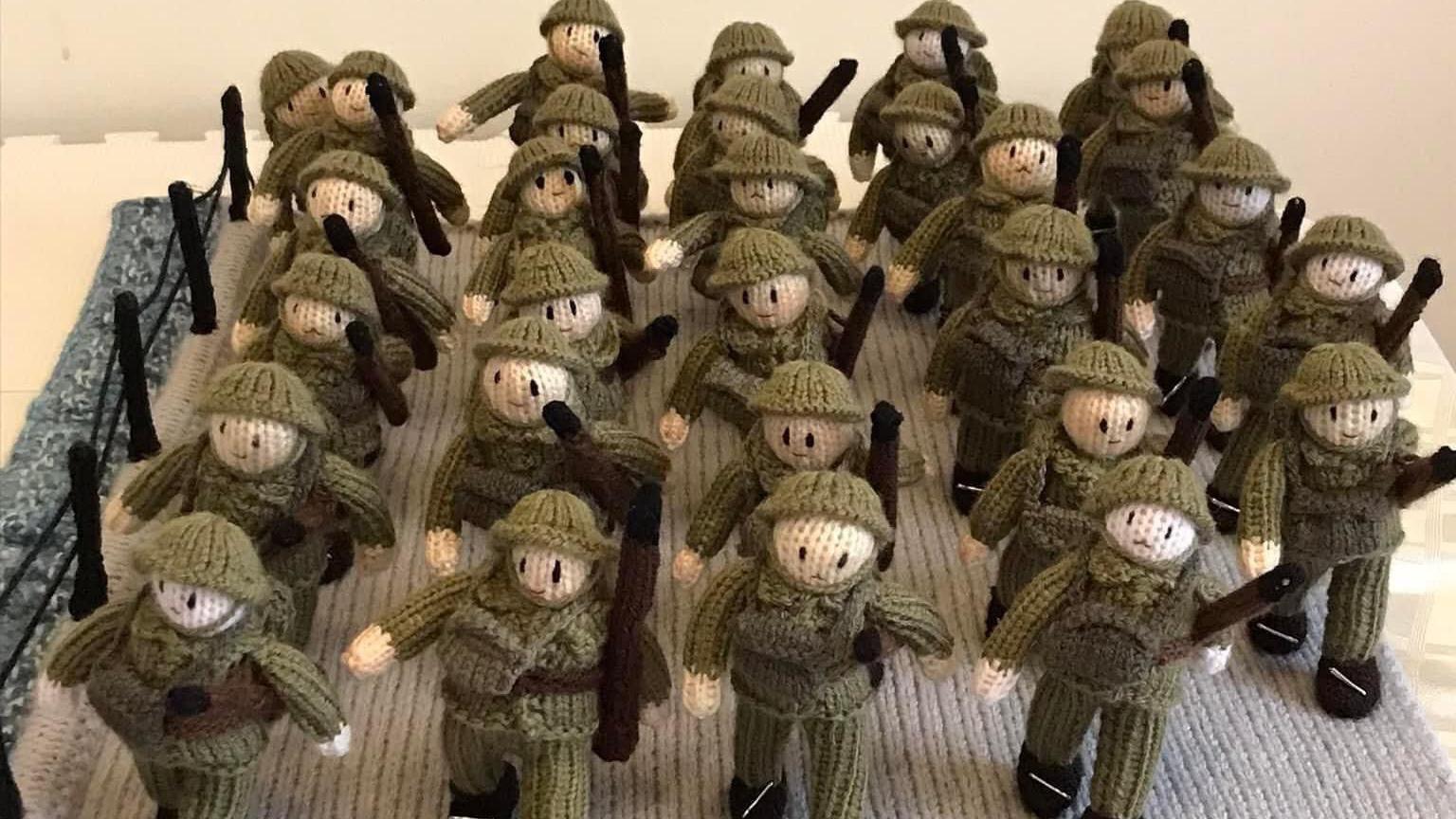 A group of 29 crocheted soldiers. They are wearing army uniforms and carrying packs and guns.