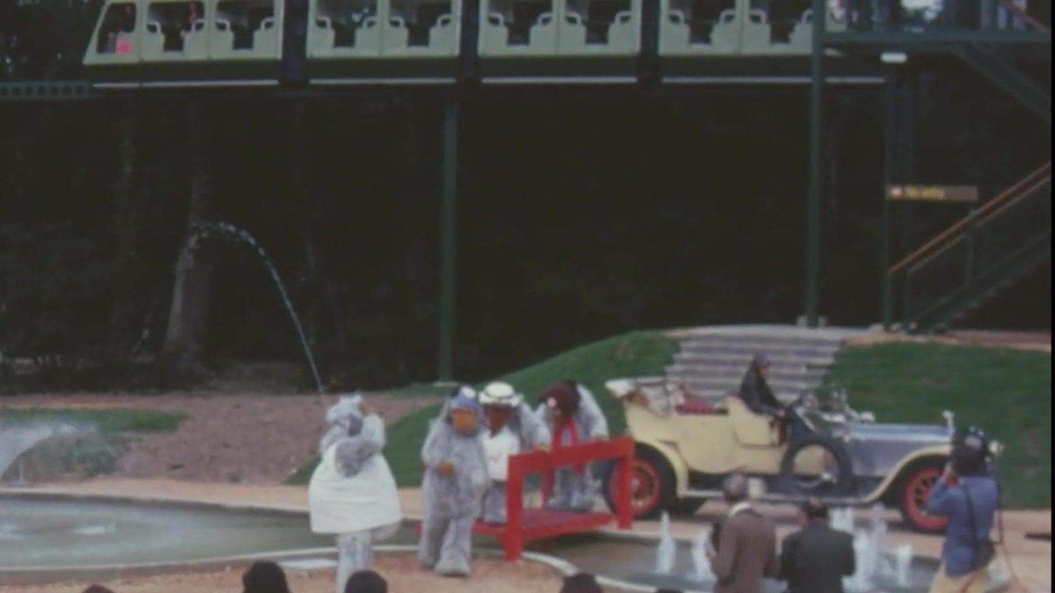 The wombles stood in front of a crowd of people, and next to the car from chitty chitty bang bang. Above them is the monorail.