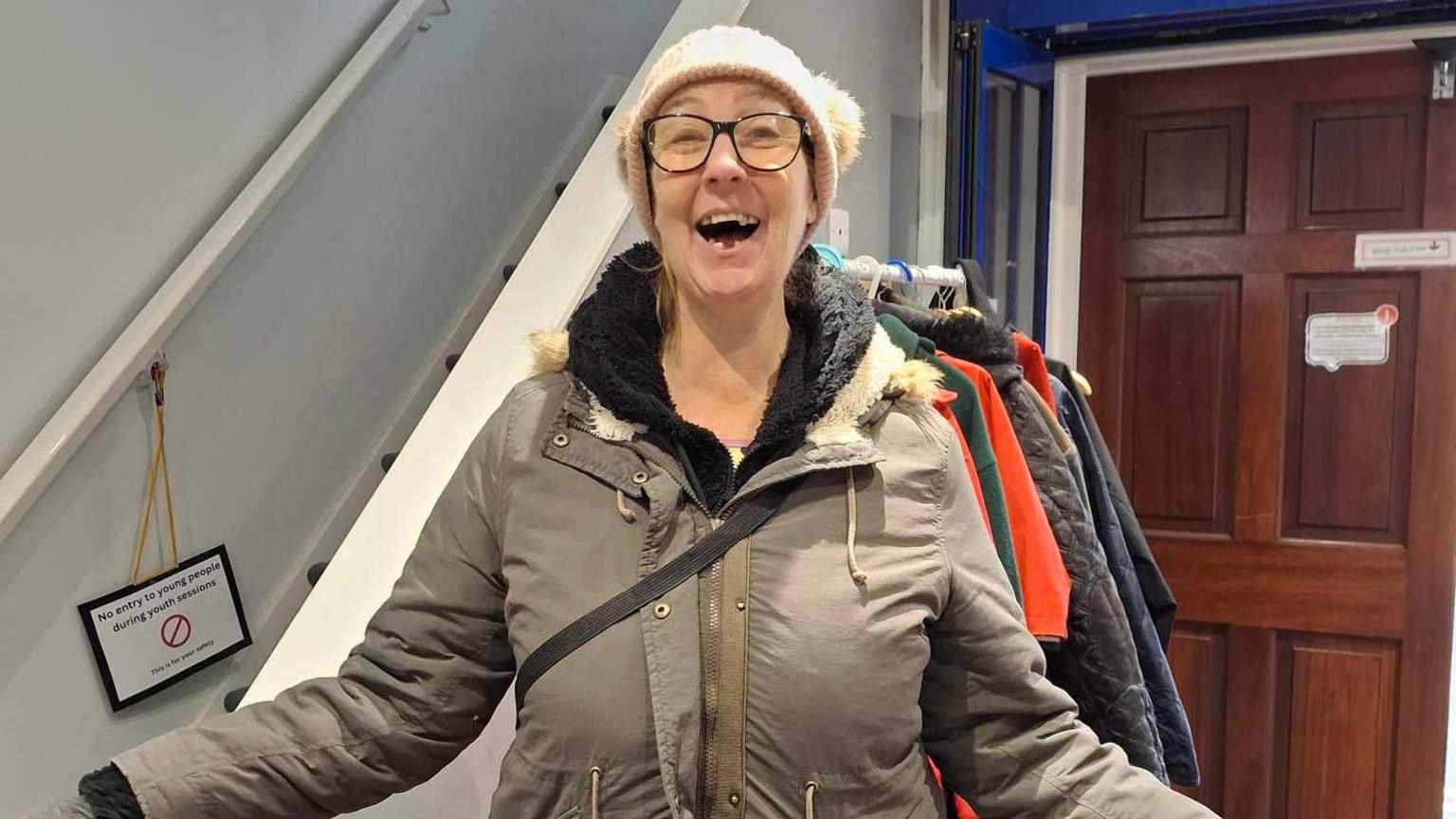 Dawn smiles at the camera whilst wearing a brown court. She has her arms outstretched and stands in front of a coat rack next to a staircase.