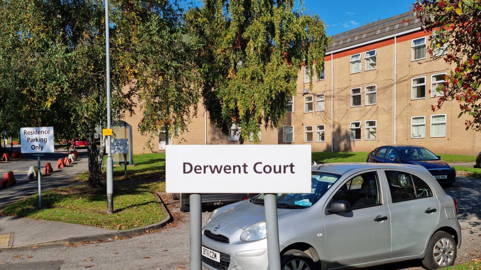 The exterior of a block of flats. A white sign infront of the flats has black writing reading 'Derwent Court'