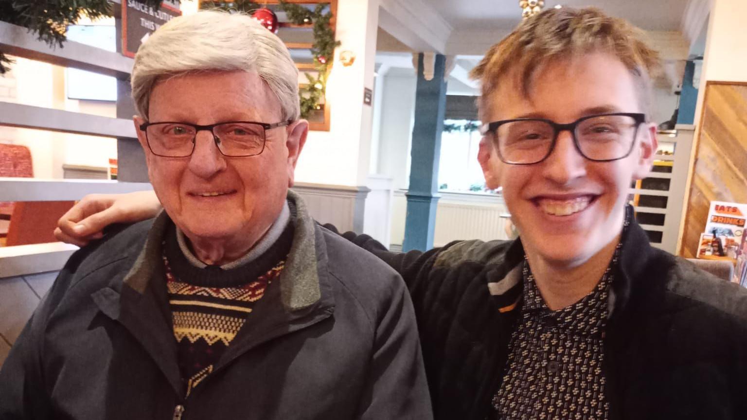Walter Nightingale sits at a table in a restaurant next to his grandson Alexander who has his arm around him. Walter has thick, short white hair and wears glasses with black rims. He wears a patterned jumper under a grey coat. Alexander has short light hair, he wears glasses with black rims. Her wears a pattered shirt under a black jacket.