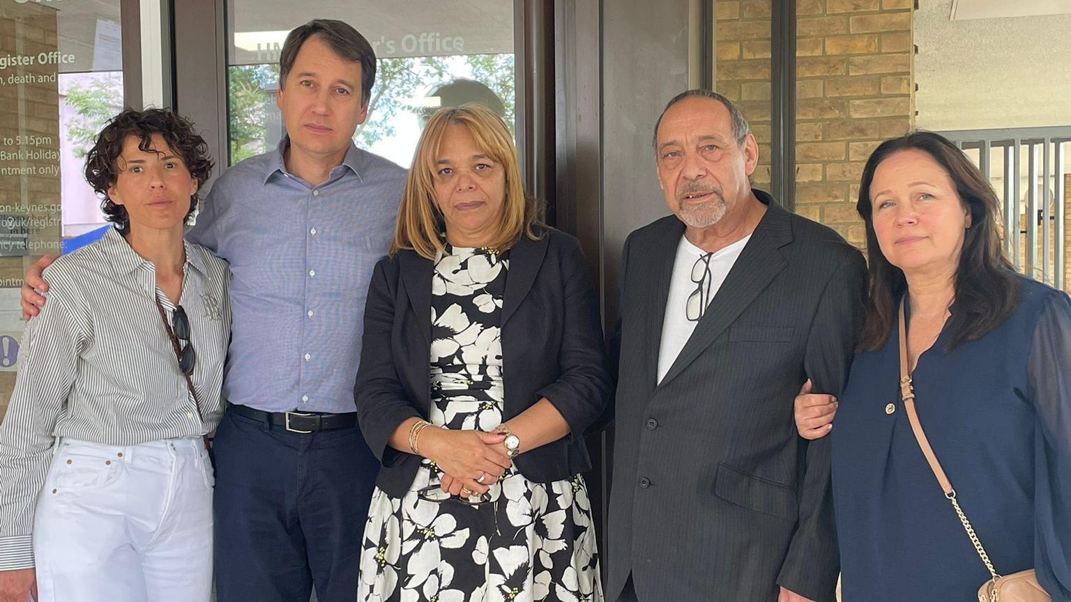 Five people stand together outside the door of the register office. They wear smart clothes and look at the camera. Aicha has a black dress with large white flowers and a blue jacket, and shoulder length blonde hair. Michael wears a white t-shirt and a grey suit jacket.
