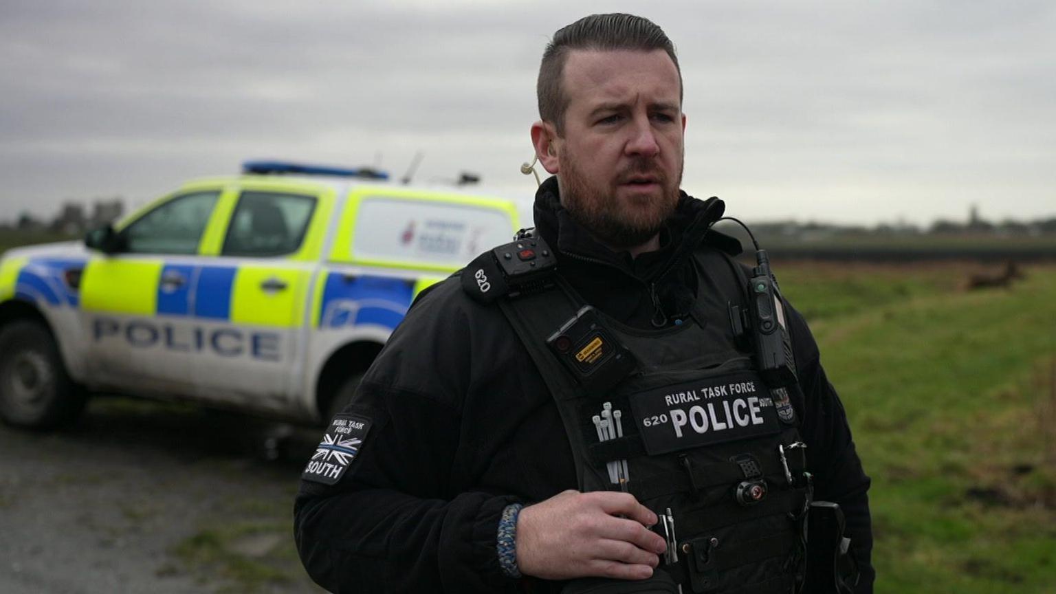 PC Gallagher speaking to the camera, surrounded by his liveried police car and green fields