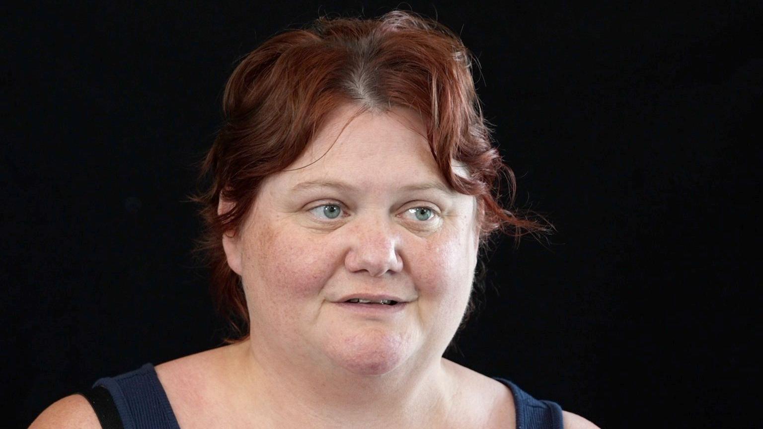 A woman with short auburn hair looks slightly off camera, mid-speech. She has blue eyes. The image is of her head and shoulders, with a black background. The straps of a dark blue vest top are on her shoulders.  