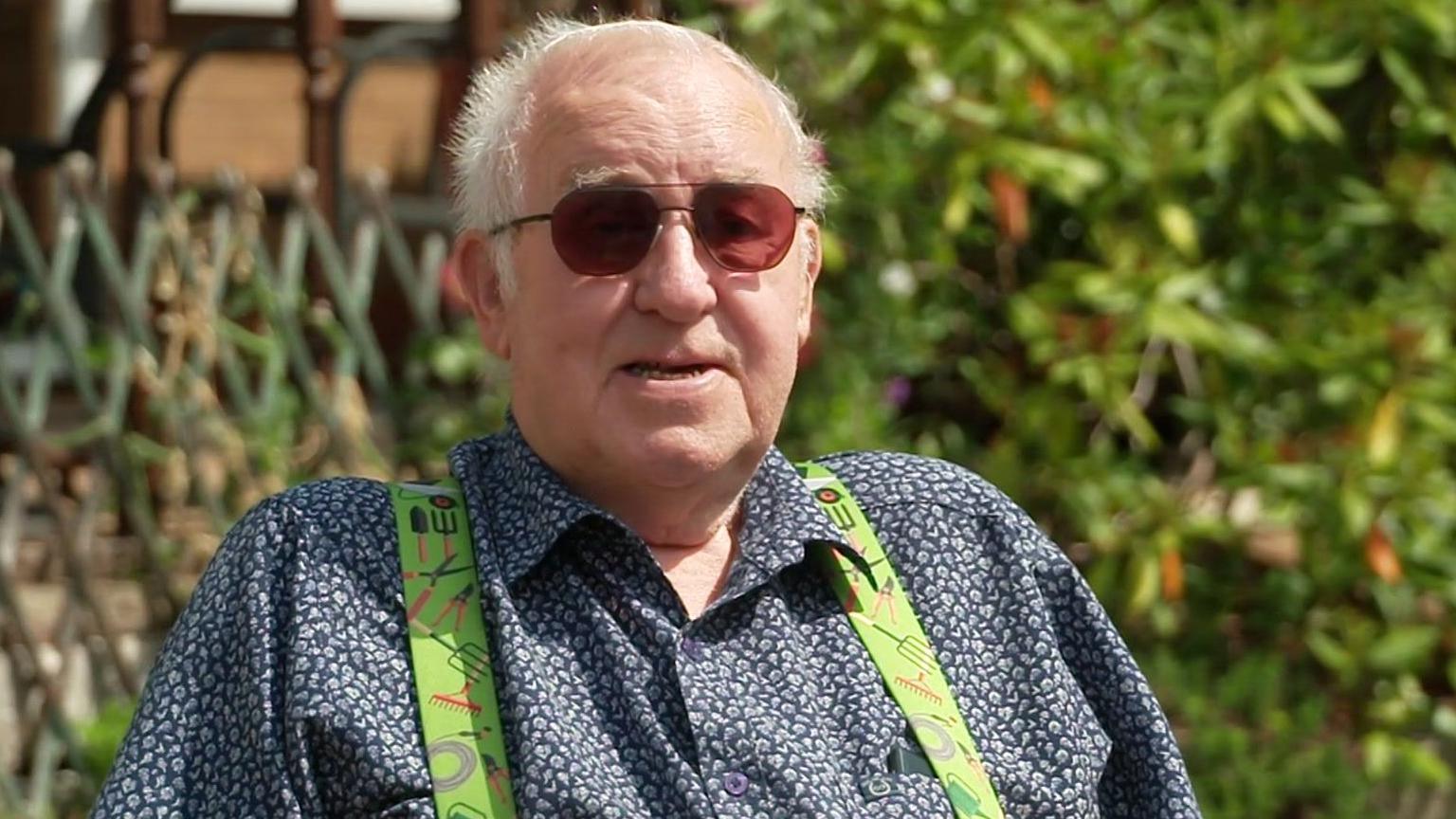 Peter Goddard has short grey hair and is wearing sunglasses, a blue shirt and green braces with gardening motifs on them. He is sat in his garden.