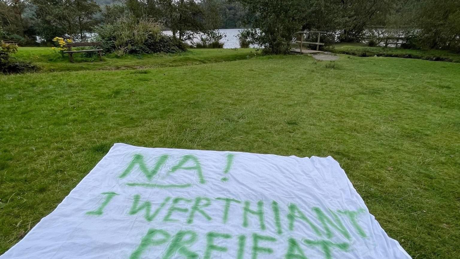 Protest banner on the grass in front of a lake with the words "Na! i werthiant preifat" in Welsh, meaning "no to a private sale"