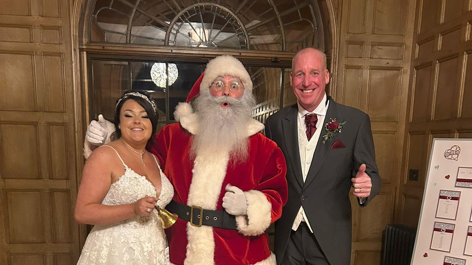 Lucy and Lee Holliday pose and smile in their wedding outfits either side of Father Christmas who arrived for their wedding reception