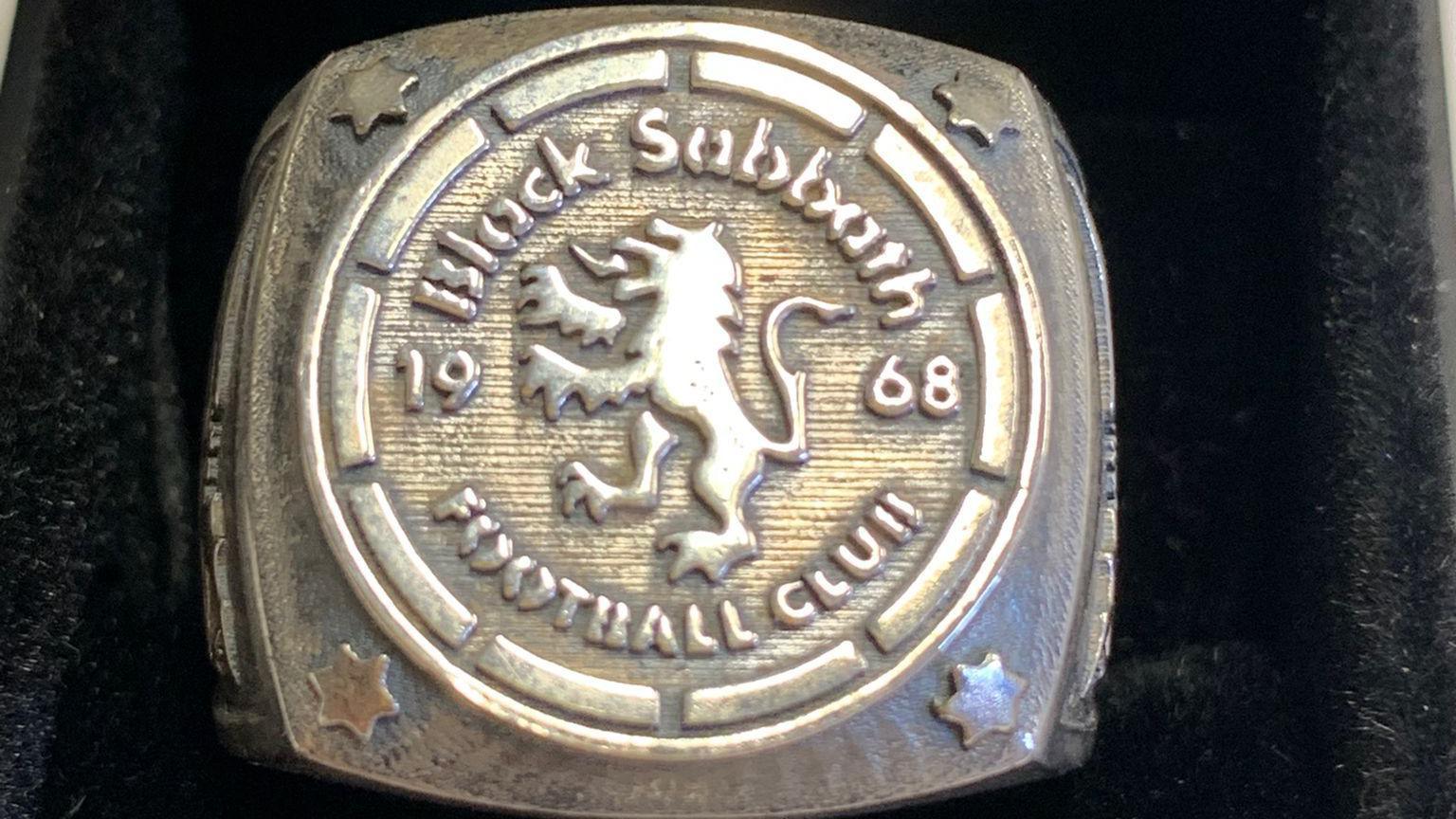 A solid silver ring is made in a curved square shape. It is decorated with a star in each corner, the football club crest and the year 1968. It is displayed in a black box.
