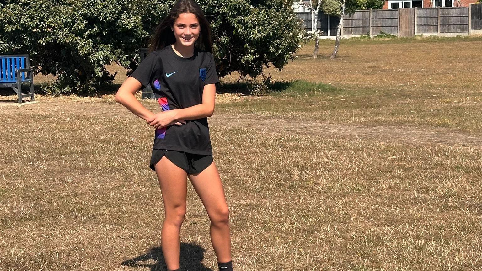 A girl standing in a brown, grassy field wearing a black England football kit
