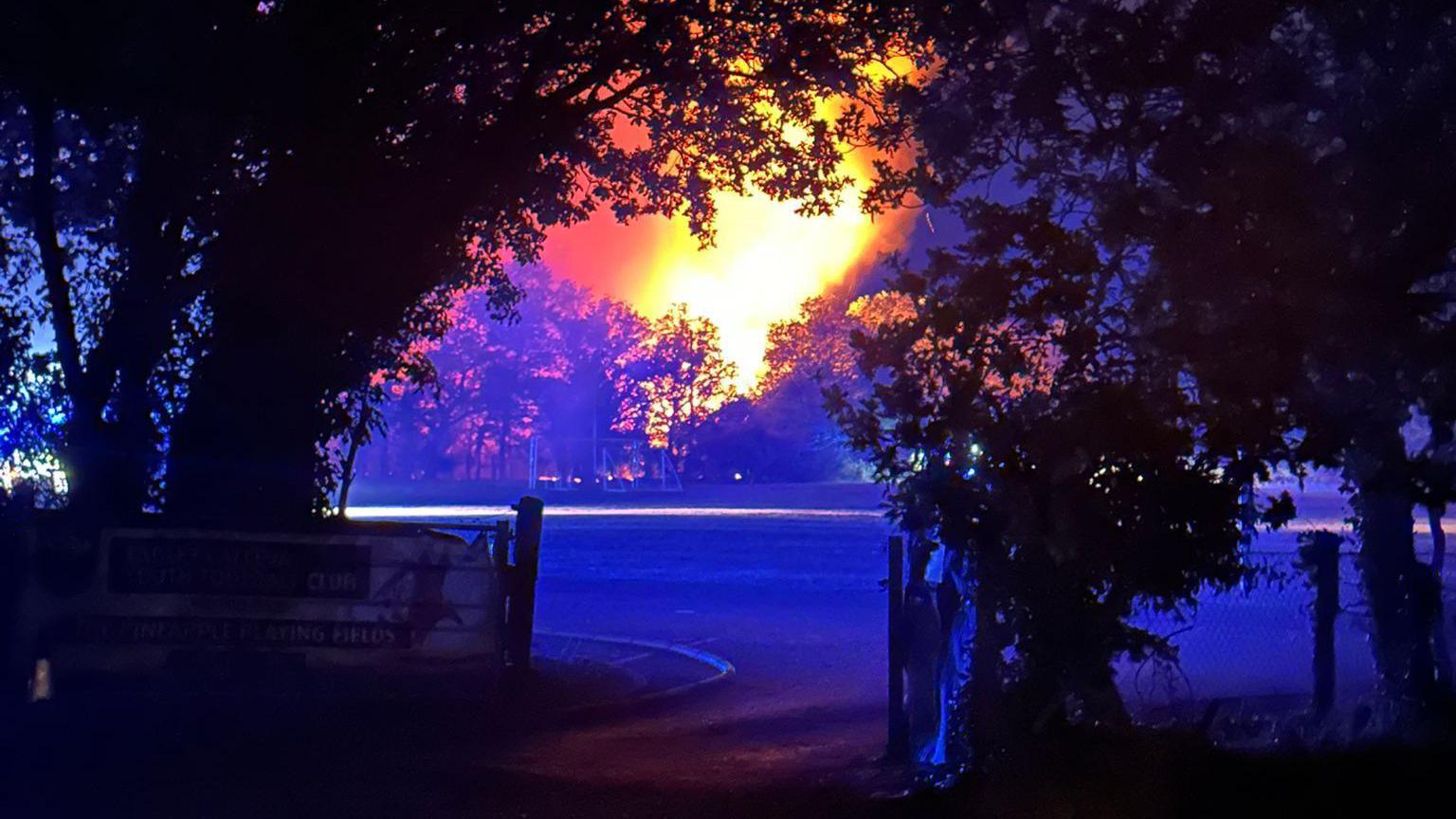 A nighttime image of the fire while it was still alight. Large flames are floating up into the air in the distance of the image, behind some trees while in the foreground of the image are the black silhoettes of some trees either side of a wooden farm gate.
