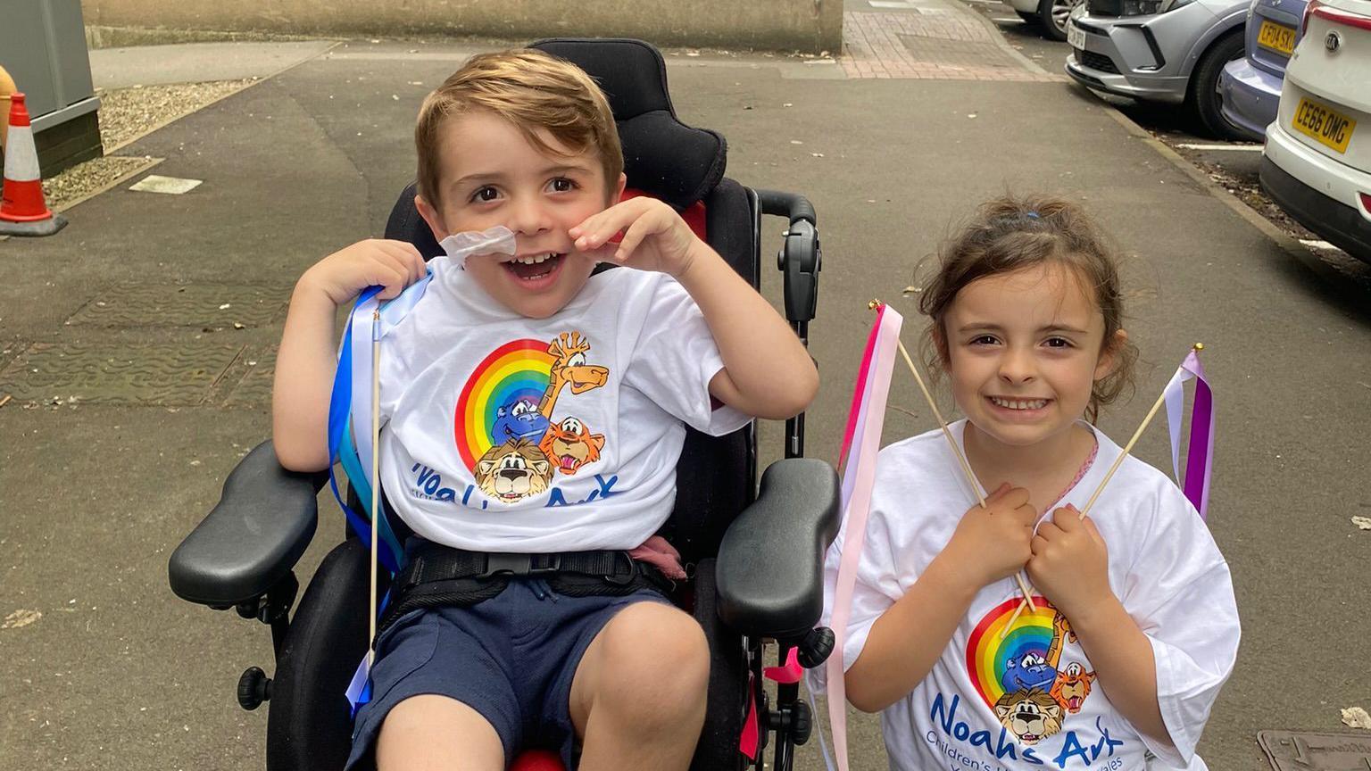 Freddie is strapped into a wheelchair with tape and a tube going into his nose. He has sandy blond hair and big brown eyes and is grinning with his hands in the air. He is wearing a white Noah's Ark shirt which matches his sister Millie who is stood next to him with streamers in her hands. She is five years old, has brown hair a big brown eyes - she too is grinning at the camera