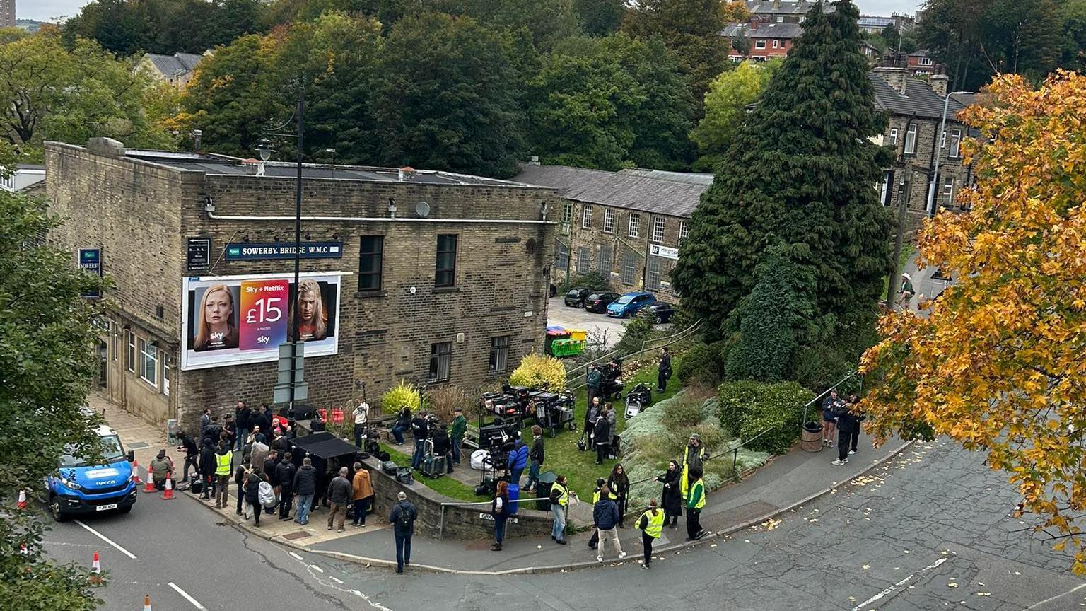 A crowd of camera crew milling around a stone building next to a row of terraced houses and trees. The building has a sign which reads 'Sowerby Bridge Working Men's Club'