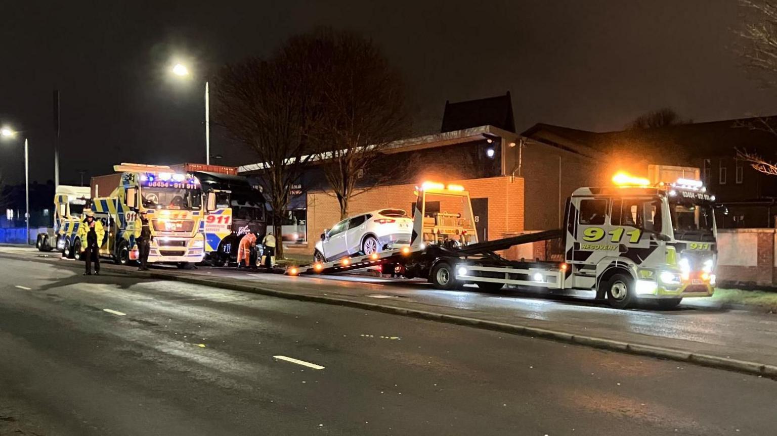 Three 911 recovery vehicles on a dual carriageway, next to a building. There is a black lorry behind one of the vehicles. A white car has been loaded onto the back of one of them. There are policeman and recovery workers at the scene. The photo has been taken at night with the scene lit by streetlights. 