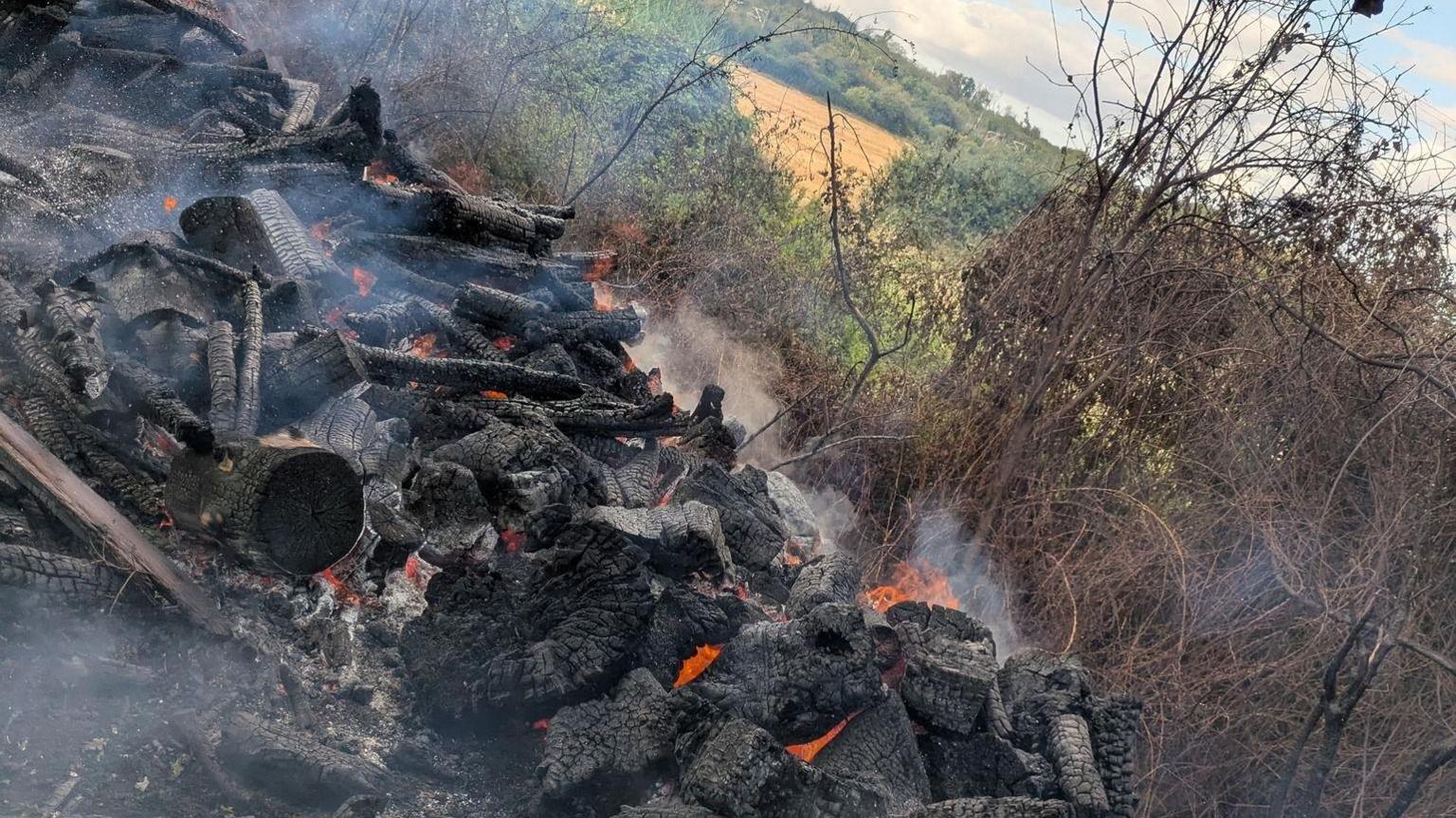 A smoking pile of charred logs and wood. Some if it is still burning with orange flames coming from the gaps in the pile.