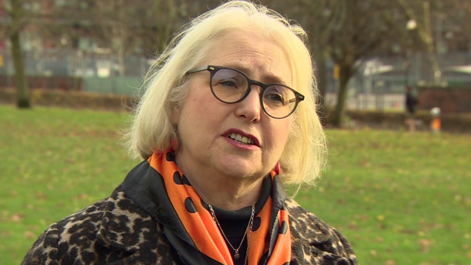 Penny Wrout seen standing in park. She is wearing glasses, a leopard print coat and a black and orange scarf.