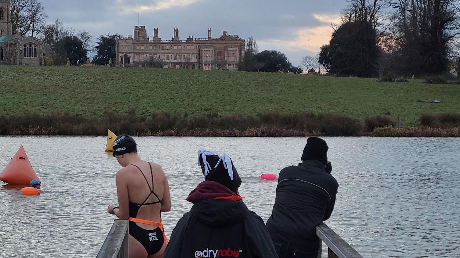 A woman in a swimming costume and cap prepares to get into a lake. Two people in navy tracksuits and hats watch on. A stately home is in the backdrop behind the lake. 