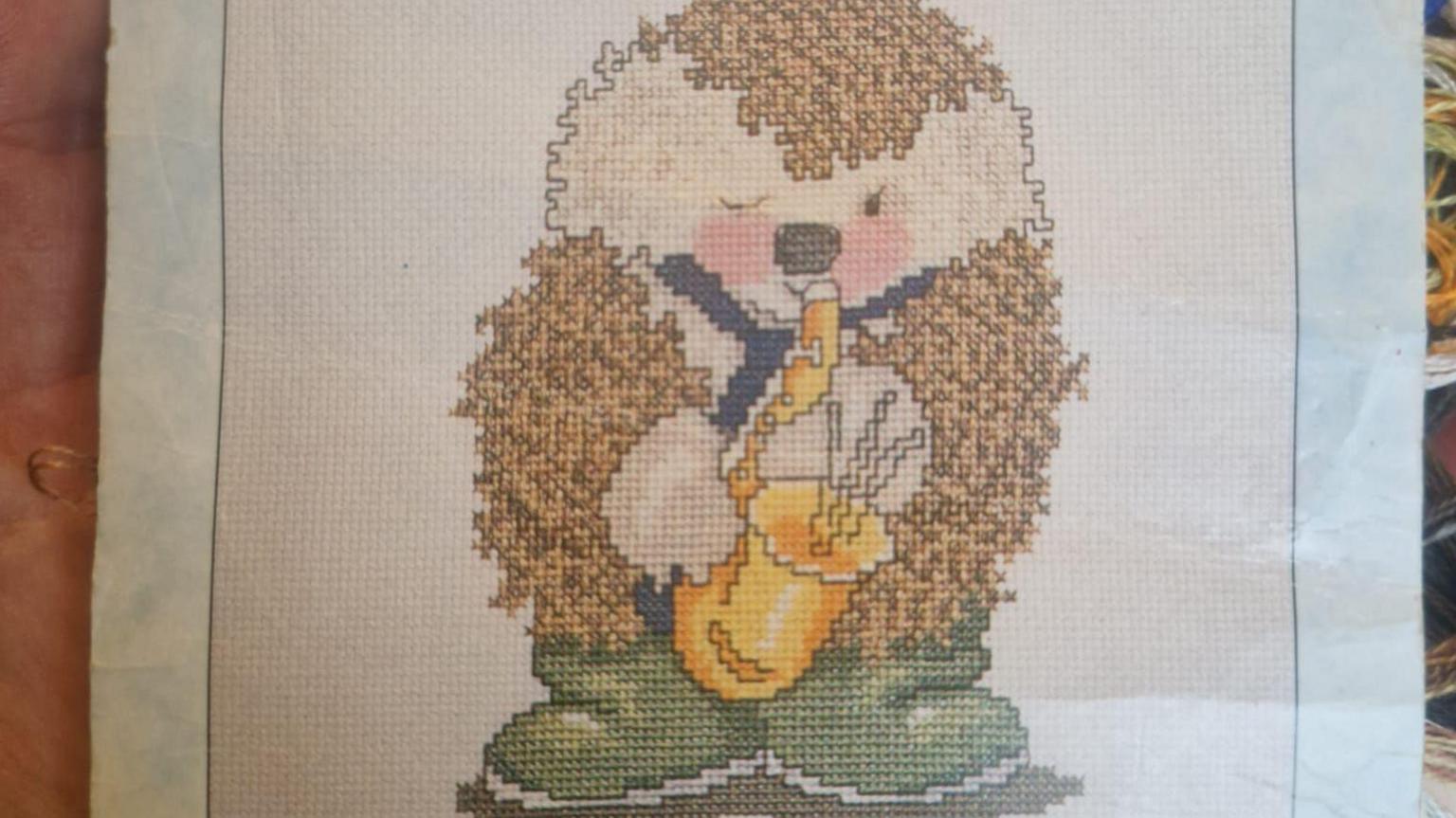 The finished cross-stitch of a hedgehog wearing a blue tie and green wellington boots playing a saxophone