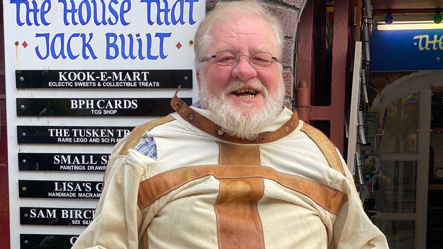 Malcolm Norton who has a white beard, glasses on and a broad teethy smile wearing the white and brown straitjacket outside the front of his shop.