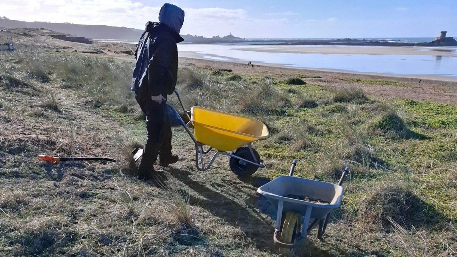 A person in a hoodie pushing a wheelbarrow on the foreshore with the sea in the background