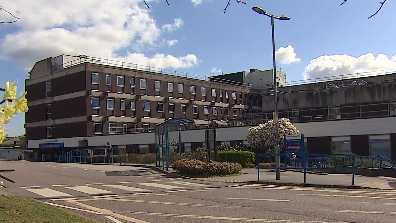 A view of outside North Devon District Hospital. The building is brown with four floors and each floor has many windows. There is a zebra crossing to the left, leading to the hospital. 