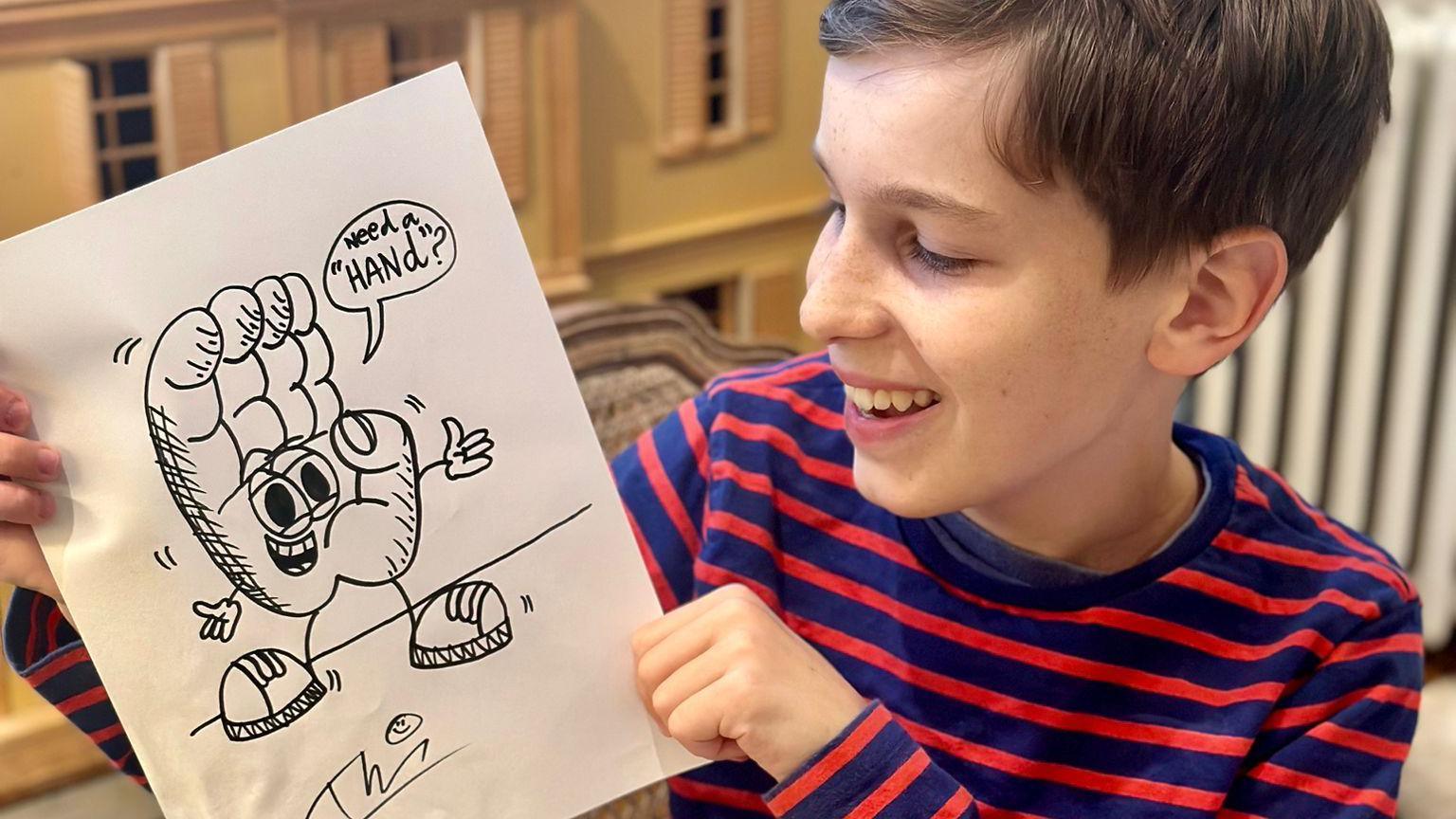 Theo, who is wearing a blue and red striped T-shirt, holding one of his drawings and smiling at it. The illustration is of a giant hand which has a face, arms and legs. A speech bubble from the giant hand says: "Need a hand?"