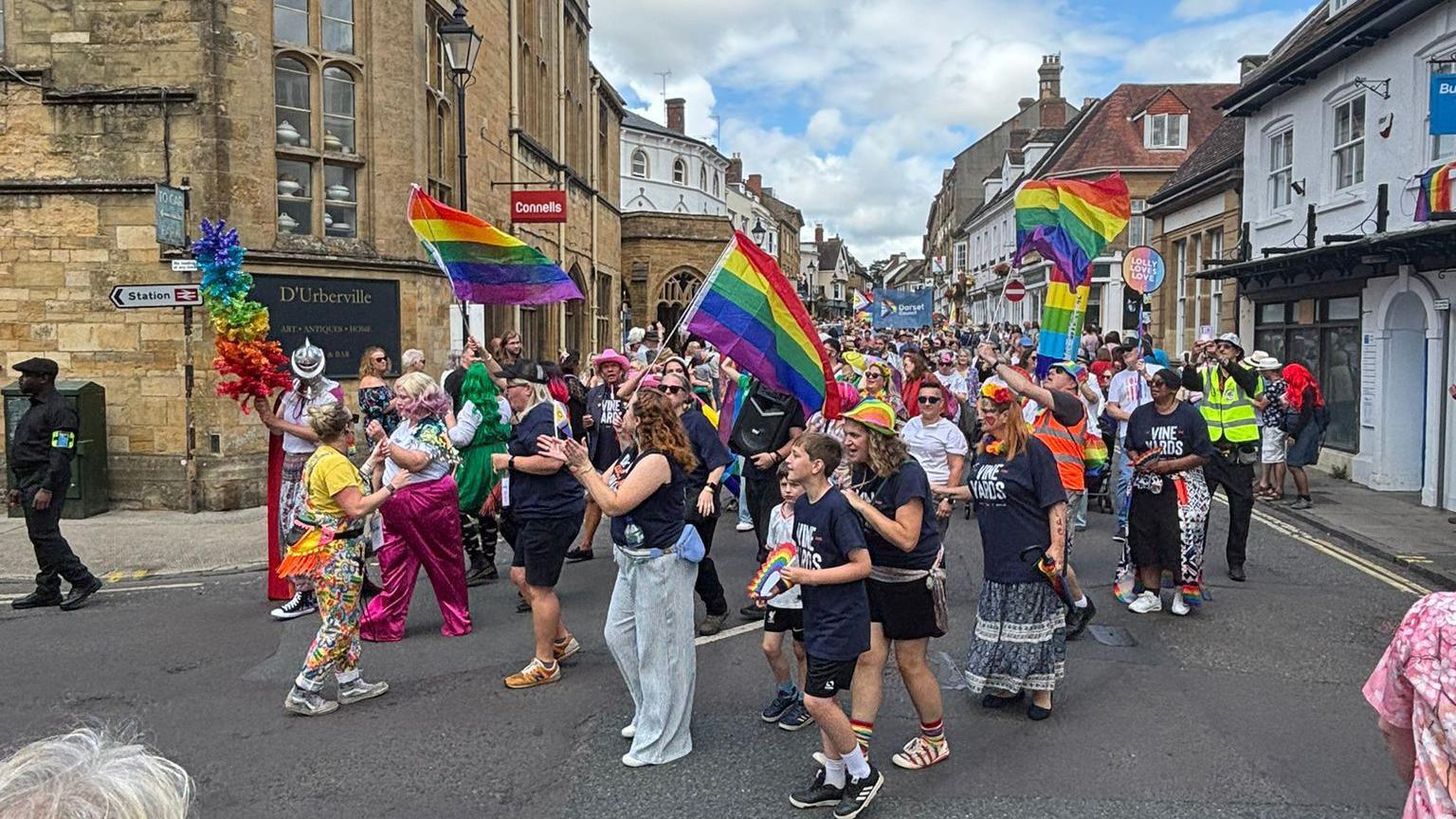 Adults and children celebrating Pride in Sherborne with a parade, waving rainbow flags as they are walking through the streets.