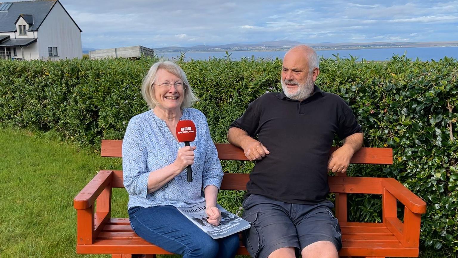 Dealbh de Christine agus Iain MacSuain agus iad nan suidhe sa ghàrradh aca agus an dithis aca a' coimhead gu math toilichte. Tha là math ann agus an t-adhar gorm agus sealladh thar na mara air an cùlaibh
