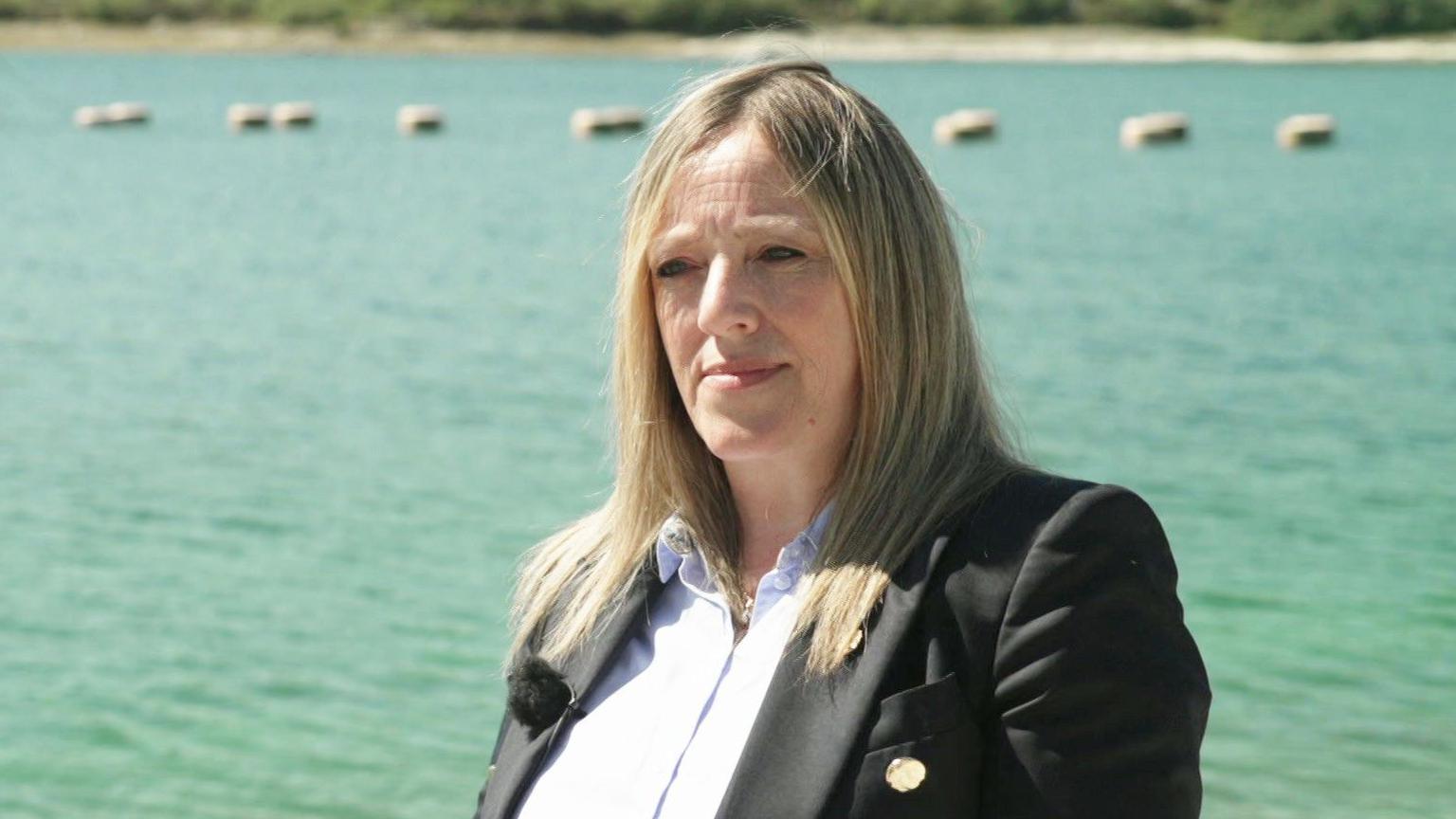 Susan Davy pictured in front of a large reservoir on a sunny day. She is wearing a light blue blouse with a dark coloured blazer. She has straight long dark blonde hair.