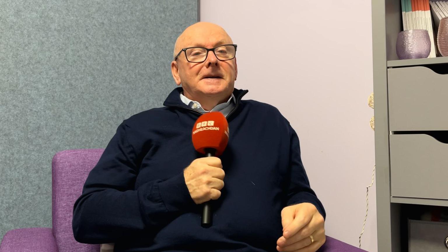 Iain MacAmhlaigh, air a robh aillse a' phròstata agus e na shuidhe agus grèim aige air microphon BBC Naidheachdan 's gàire air aodann. 